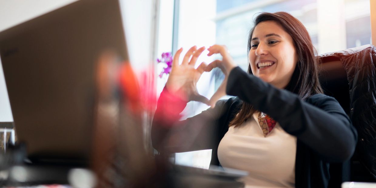 Happy woman making a heart shape with her hands in front of a laptop.