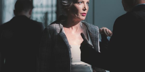 Mature woman in a checkered suit engaged in a serious conversation with a man in a dimly lit space.