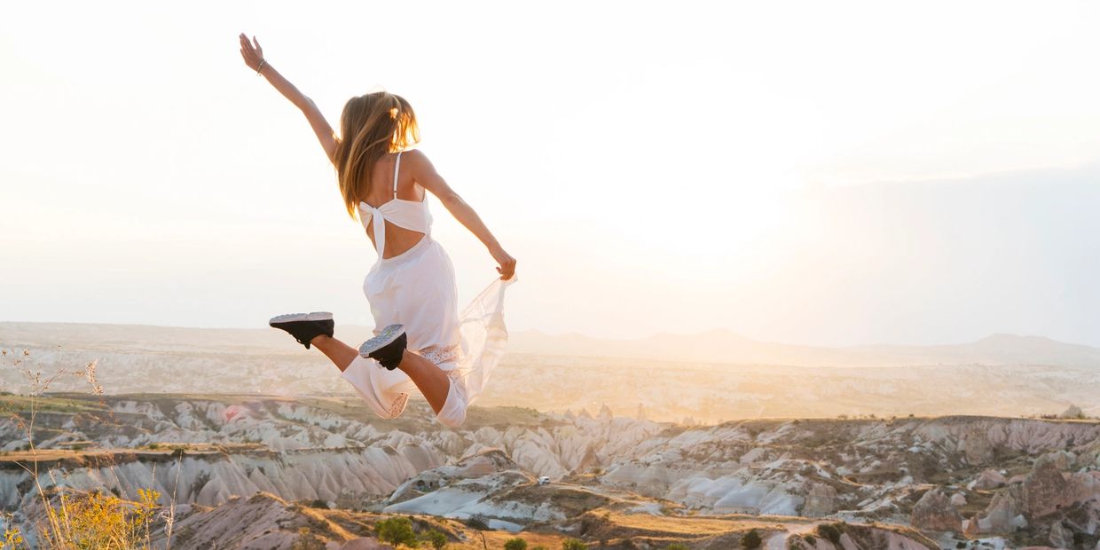 Woman in white dress joyfully jumps over rocky landscape at sunset.