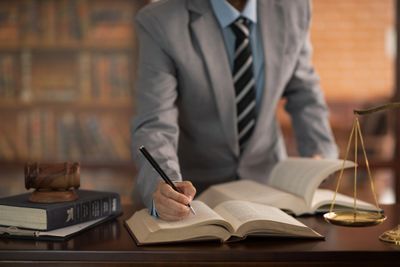 Lawyer in suit writing notes with legal books, gavel, and scales on desk.
