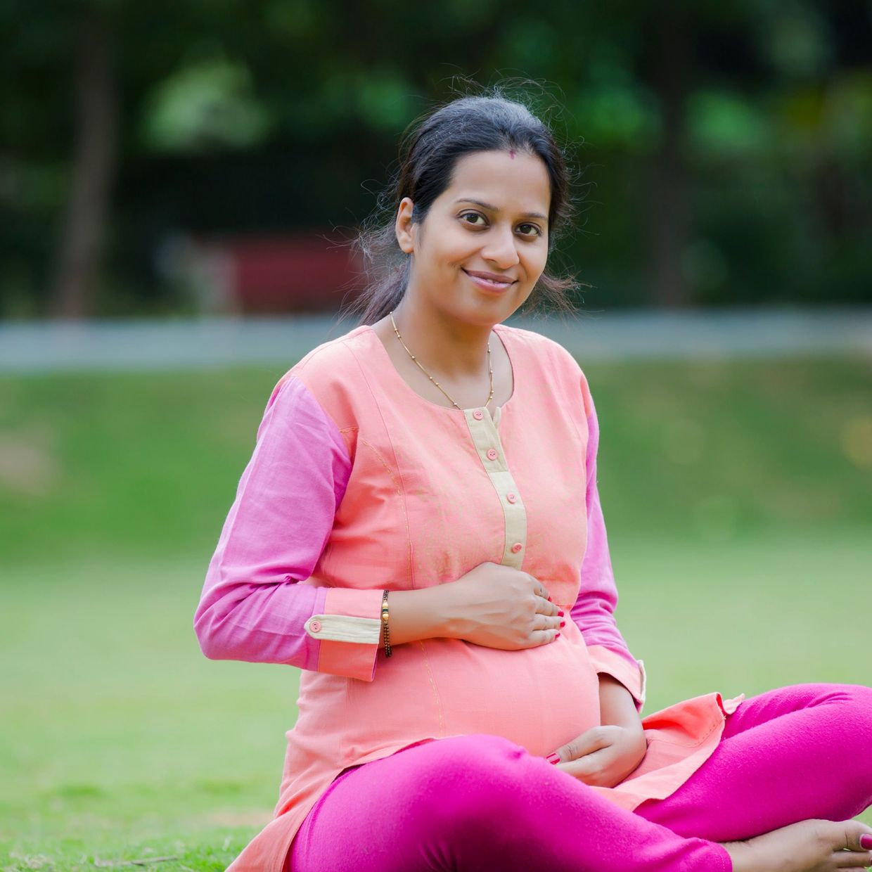 Pregnant woman sitting on grass smiling, holding her belly.