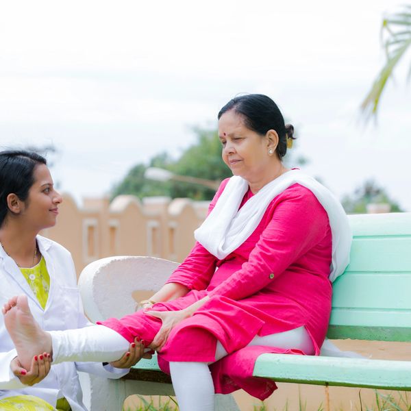 A female doctor examining an elderly woman's foot on a bench outdoors.