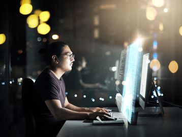 Man working late on computer with code hologram in dark office.