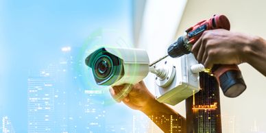 Hands installing a security camera with a drill against a cityscape background.