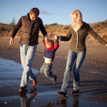 Parents joyfully swing their child while walking along the beach at sunset.