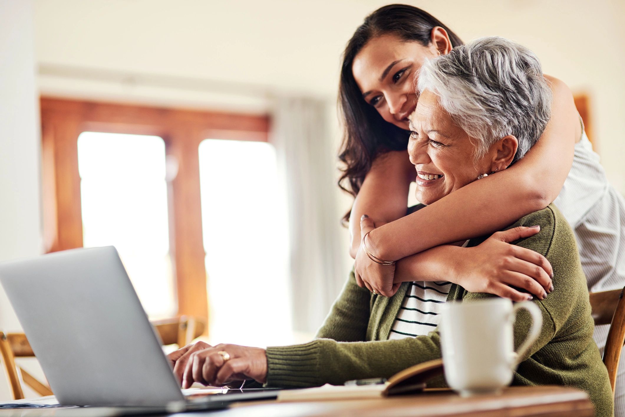 A young woman hugs an older woman using a laptop, both smiling warmly.