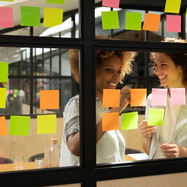 Two women collaborating with colorful sticky notes on a glass wall in an office.