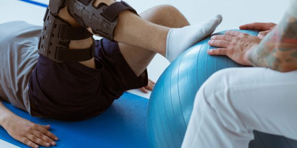 Person undergoing knee rehab with leg brace and exercise ball.