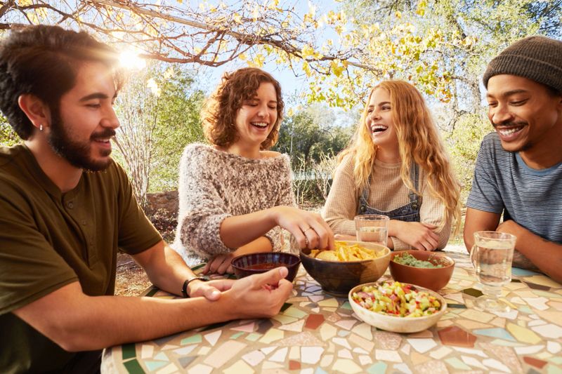Group of diverse young friends laughing and eating snacks outdoors at a table on a farm