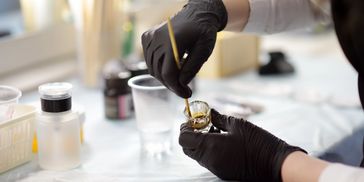 Person wearing black gloves applies substance with a brush to a small container.