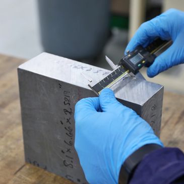Person measuring a metal block with digital calipers while wearing blue gloves.