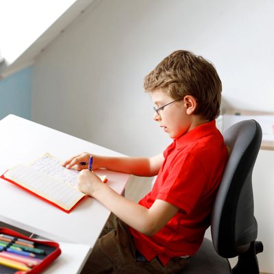 Young boy in red shirt focused on writing at a bright desk.