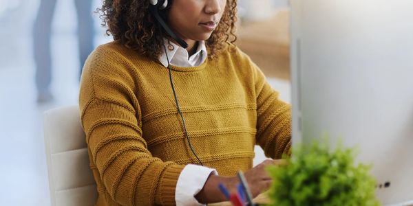 Woman in a mustard sweater working at a computer with a headset on.