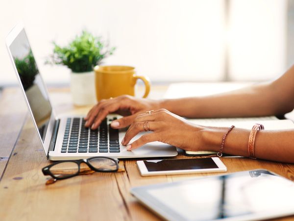 Person typing on laptop with coffee, glasses, phone, and tablet on wooden desk.