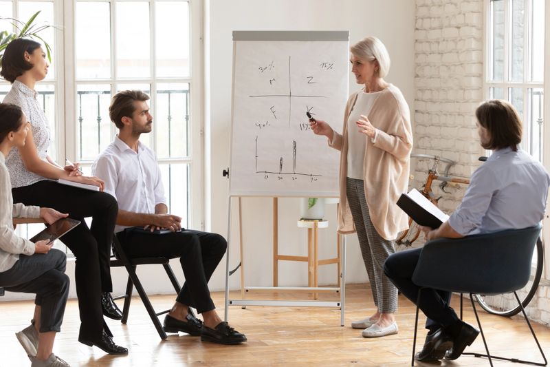 Skilled old female coach teacher mentor manager give flip chart presentation training staff group during business workshop, middle aged businesswoman trainer teaching young employees at team meeting
