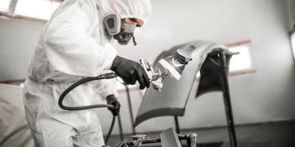 Person in Schutzanzug sprüht Farbe auf Autoteile.