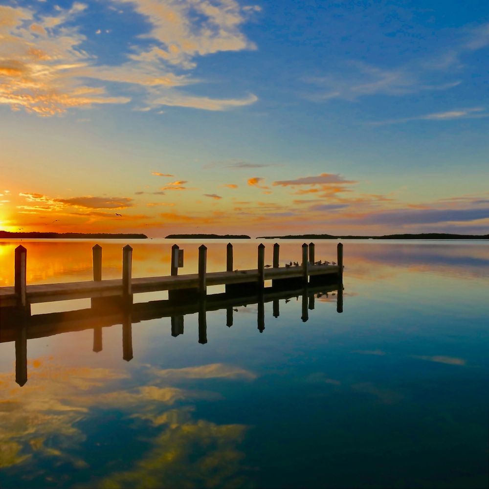 Sunset over a calm lake with a wooden pier and birds perched on it.