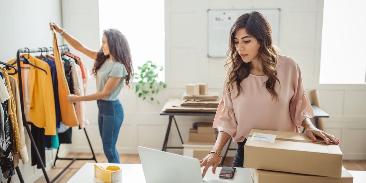 Two women working in a clothing and packaging workspace with a laptop and boxes.