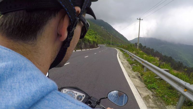 POV: Sitting behind a male tourist riding a motorbike across the stunning Hai Van pass. Scenic view of the lush tropical forest as young man drives you on his motorcycle along the picturesque road.