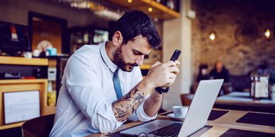 Man with tattoos concentrates on his phone beside a laptop in a cozy cafe.