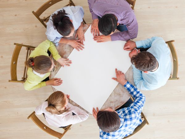 Six people sitting around a round table looking at a blank whiteboard.