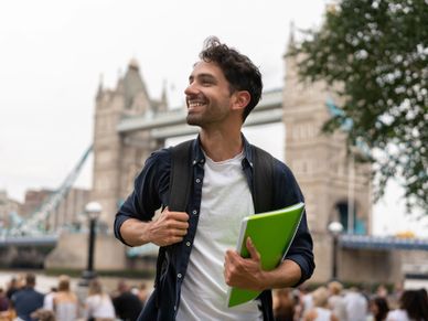 A smiling young man holding a green folder in front of Tower Bridge, London.