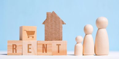 Wooden blocks spelling RENT with a cardboard house and wooden family figures.