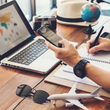 Person holding phone, writing notes near laptop and travel items.