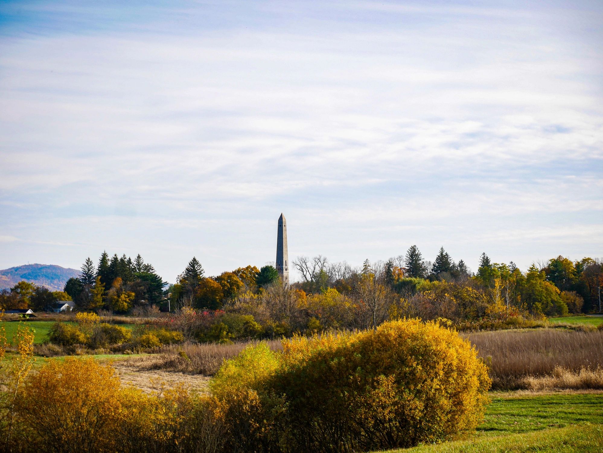 A tall obelisk monument surrounded by autumn trees and fields under a cloudy sky.