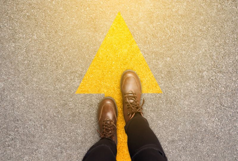 Feet and arrows on road background in starting line beginning idea. Top view. Woman in leather ankle boots on pathway with yellow direction arrow symbol. Moving forward, new start and success concept.