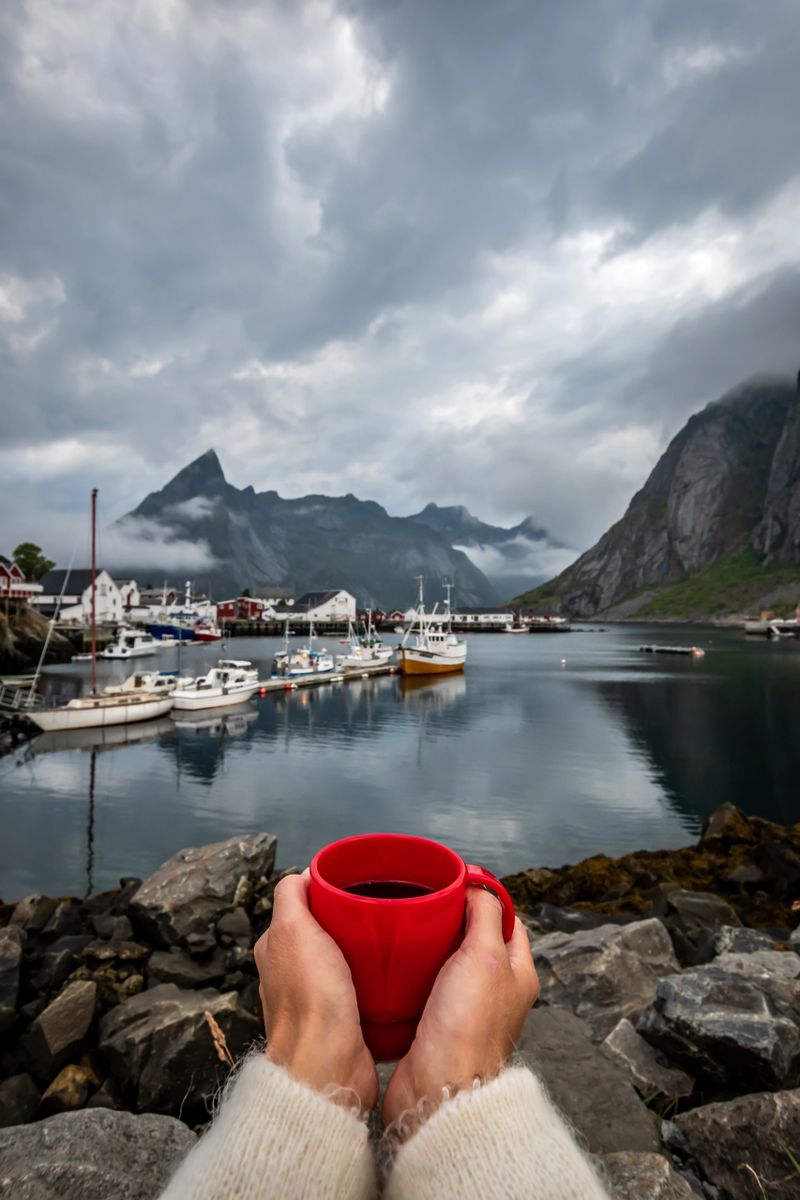 POV photo of a woman having a coffee in the fishing village of Hamnøy in the Lofoten Islands with mountain range and cloudy sky in background during summer.