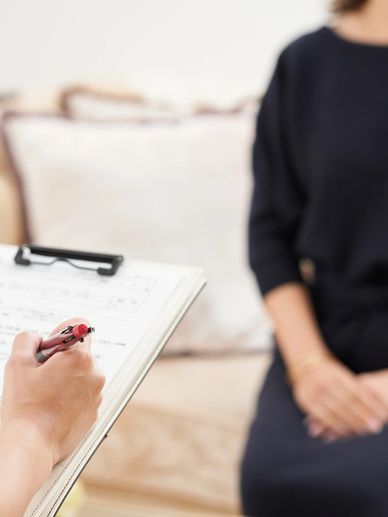 A person taking notes on a clipboard while another sits nearby, possibly in a counseling session.