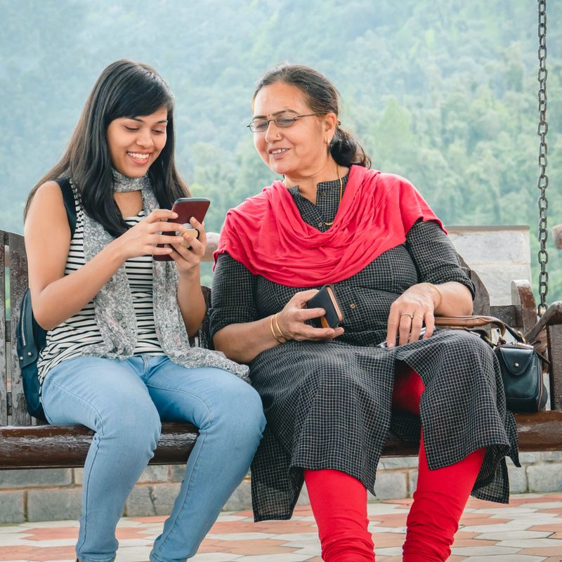 An Asian/Indian beautiful daughter sits on a porch swing and teaches the smartphone operating to her senior retired mother at a resort in the mountains of Himachal Pradesh.
