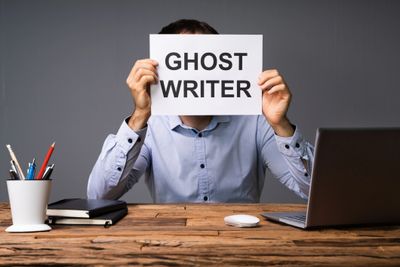 Person holding a 'GHOST WRITER' sign in front of their face at a desk.