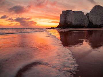 Sunset over a beach with cliffs and gentle waves reflecting vibrant colors.