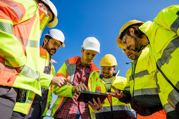 Construction workers in safety gear discussing plans on a tablet under clear blue sky.
