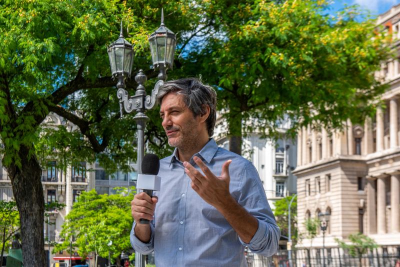Male mature reporter talking and holding a microphone in a public park during a sunny day at Buenos Aires, Argentina. At the background the facade of the Palace of Justice.