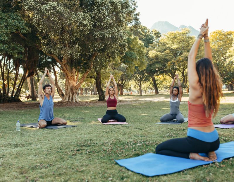 Yoga teacher leading her students through their poses while sitting on exercise mats outside in a park on a sunny day