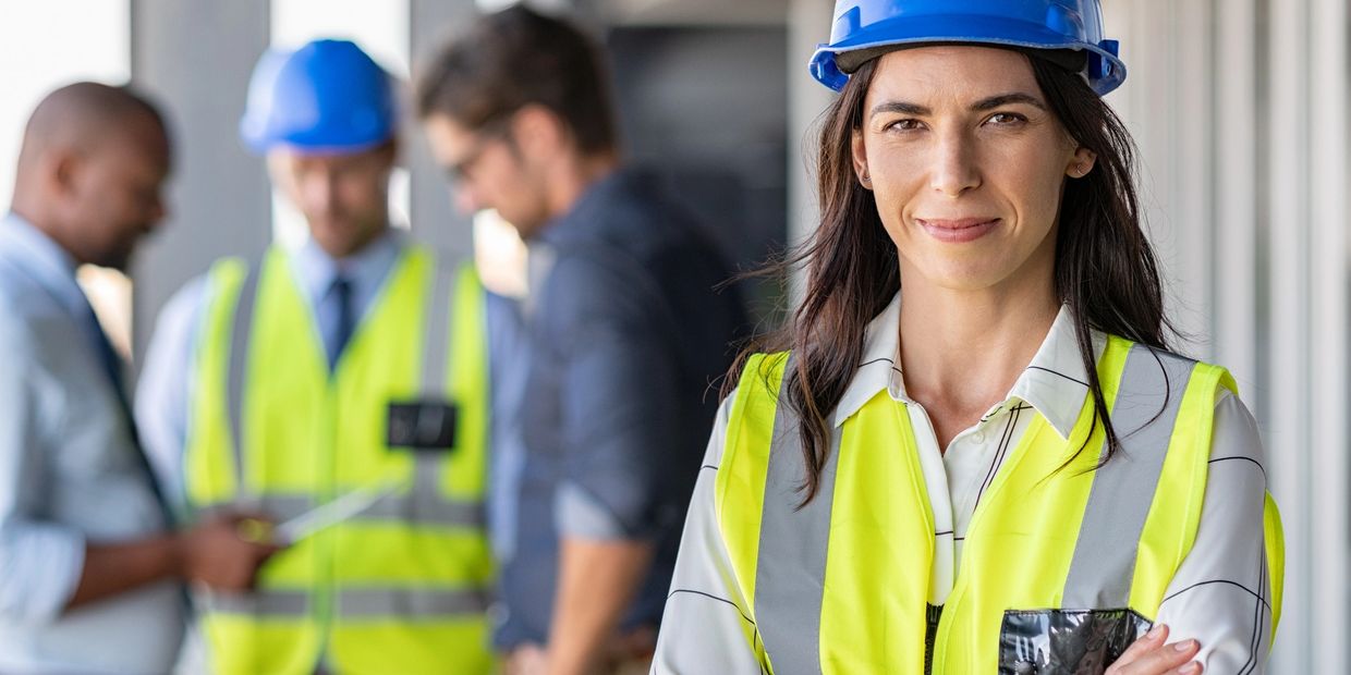 Female engineer in safety gear stands confidently with team in background.