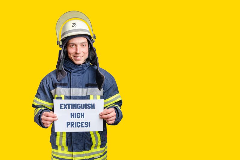 young smiling firefighter wearing uniform and helmet looking to the camera and holding paper sheet with inscription extinguish high prices in hands. isolated on yellow background