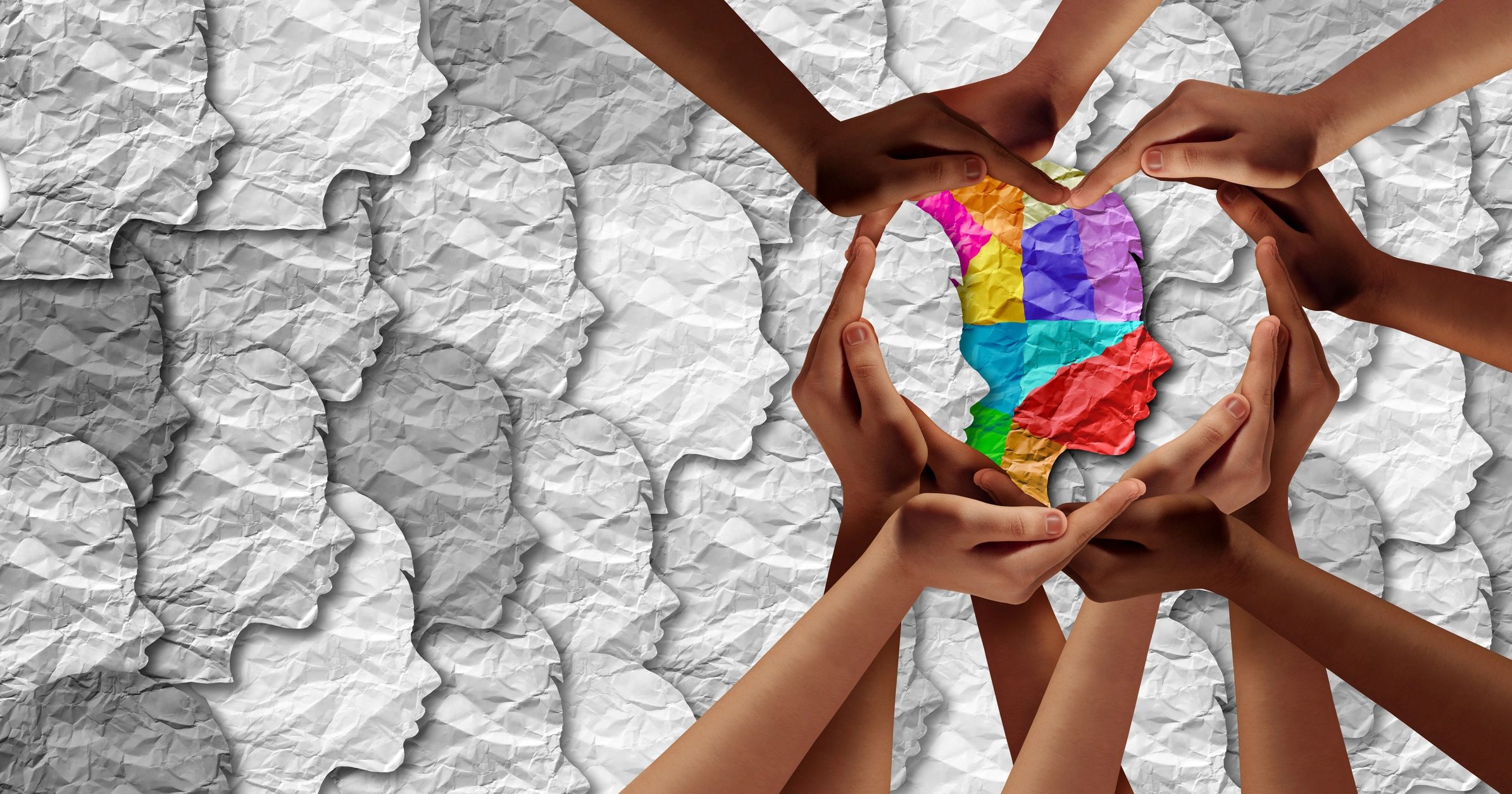 Diverse hands forming a heart around a colorful paper face among gray paper faces.