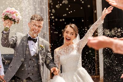 Newlywed couple joyfully celebrates as guests throw confetti outside a building.