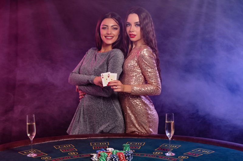 Two beautiful brunette women are playing poker at casino, hugging, holding cards, posing at a table with stacks of chips, champagne on it. Black, smoke background, colorful backlights. Close-up.