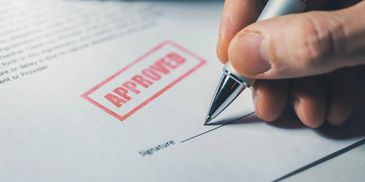 A hand signing a document stamped with 'APPROVED'.
