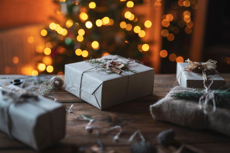 Close up of Christmas presents wrapped in natural materials on wooden table with blurred light bulbs on background.