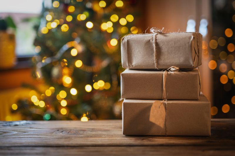 Close up of Christmas presents wrapped in natural materials on wooden table with blurred light bulbs on background.