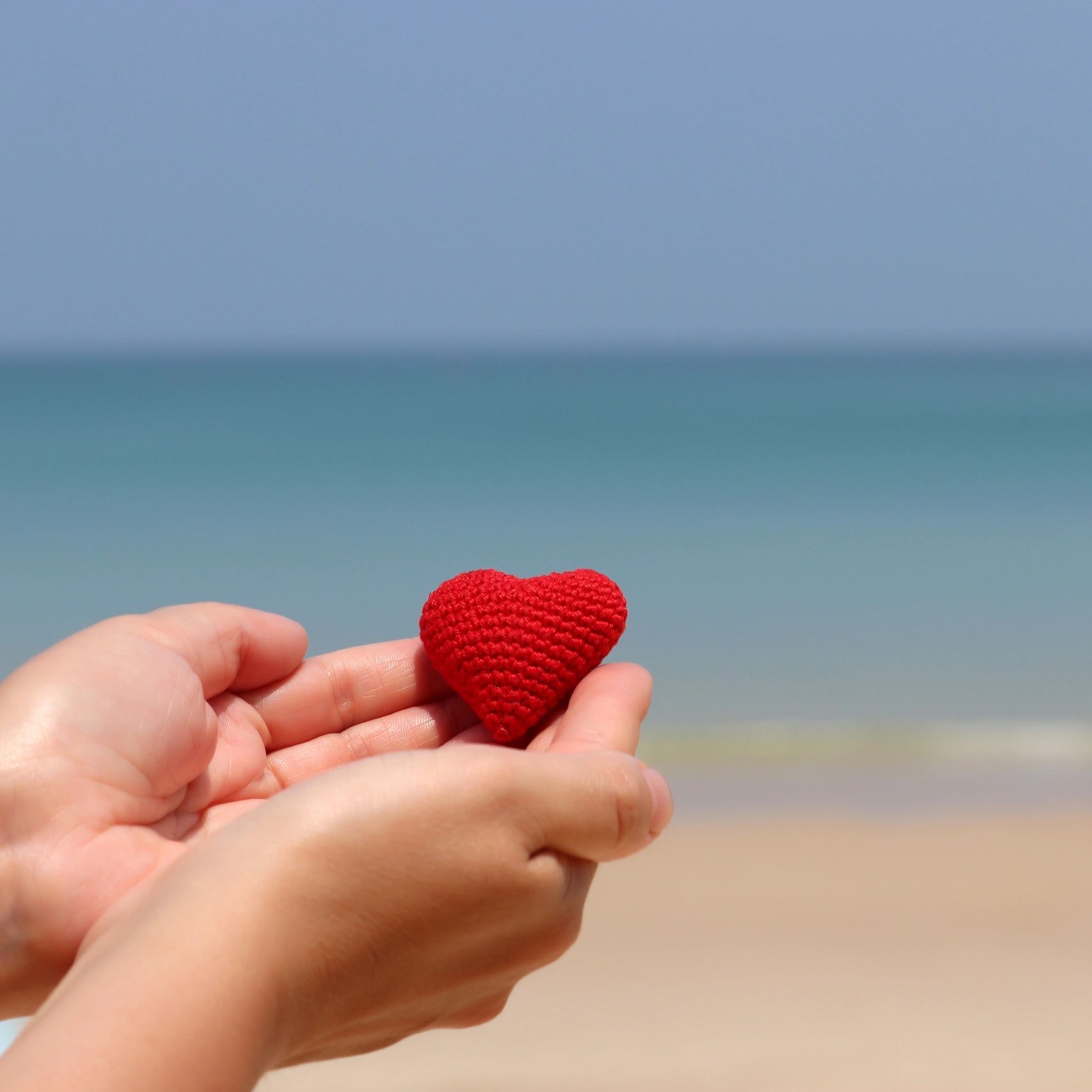 Hands holding a red knitted heart with a beach background.