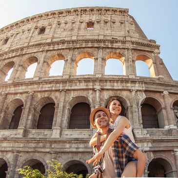 A man gives a girl a piggyback ride in front of the Colosseum in Rome on a sunny day.
