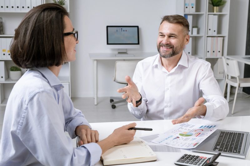 Happy mature broker and his young female colleague discussing financial documents in office