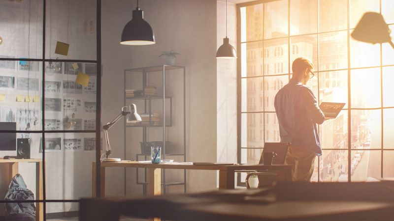 Thoughtful Young Professional Designer Sits at His Desk and Looks onto Big City out of His Windows, Contemplating Next Steps in Finishing Project, Writes Down His Ideas. Handsome Professional Working